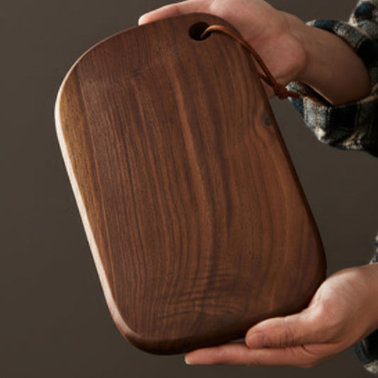 A person holds a dark brown wooden cutting board with rounded corners and a hanging loop.