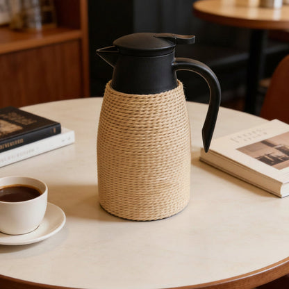 A black insulated coffee carafe with a beige woven straw sleeve sits on a round light wood table in a cafe setting, alongside a cup of coffee, books, and other table decor.
